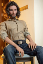 A mannequin with long hair and a beard, wearing a tan button-up shirt and jeans, sits on a chair.