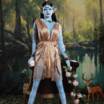Blue-skinned fantasy character with gold dress and horns, holding roses, seated in a forest setting.