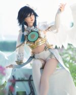 A person in a white and gold costume with blue hair and large white wings sits on a stone surface outdoors.